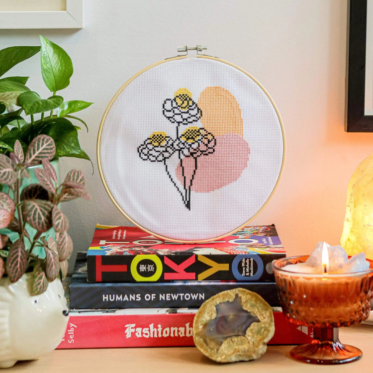 Craft Club Co BLOOM Cross Stitch Kit. The design is shown sitting on a stack of books with a candle and pot plants around it. The design is three line art flowers, with organic pink and orange shapes behind it. There is also yellow spotches on the flowers.
