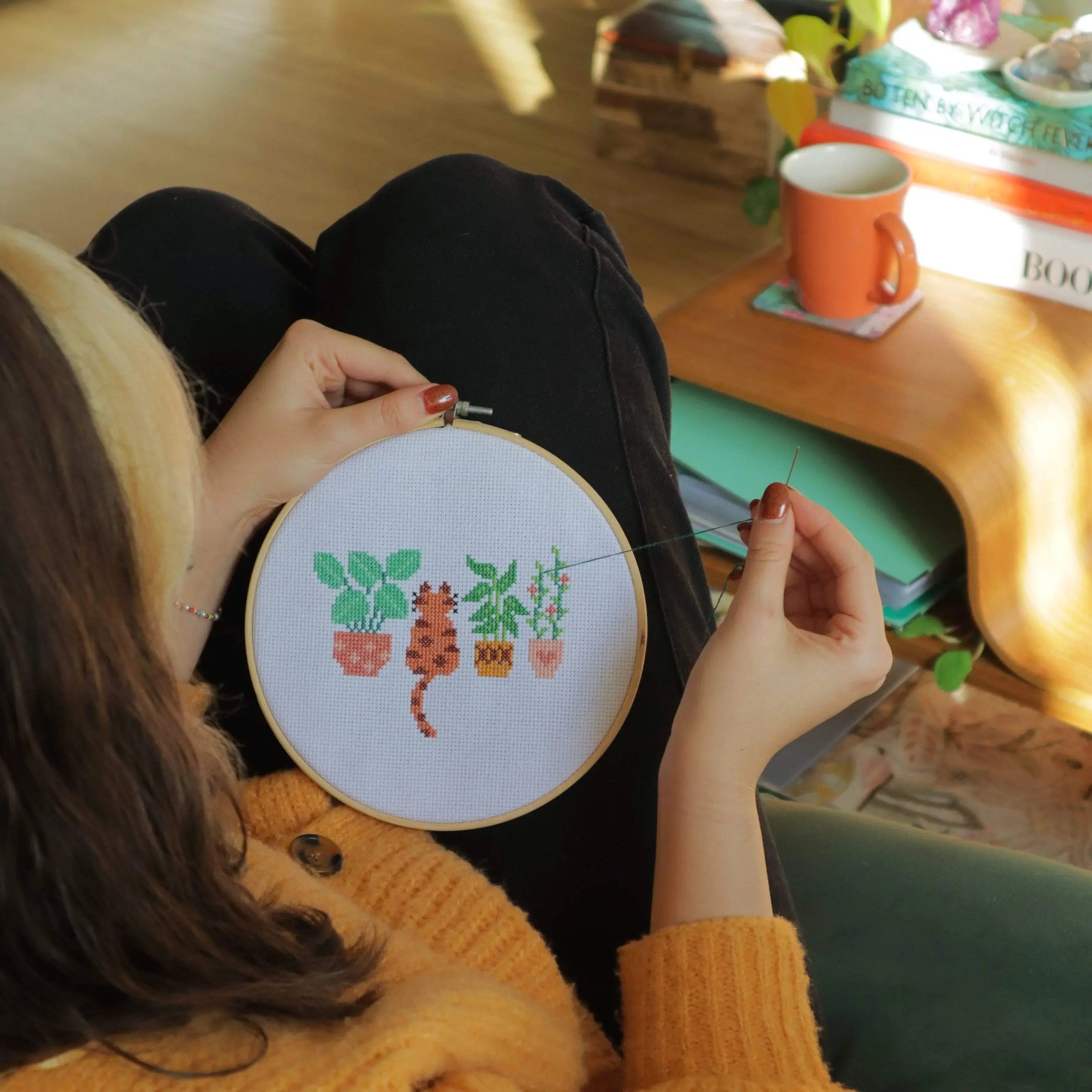 Craft Club Co CAT AMONGST THE POT PLANTS Cross Stitch Kit. Looking over someone's shoulder, the design is being stitched. The person is sitting at a coffee table with a cup of tea, and is holding the threaded needle as they work.