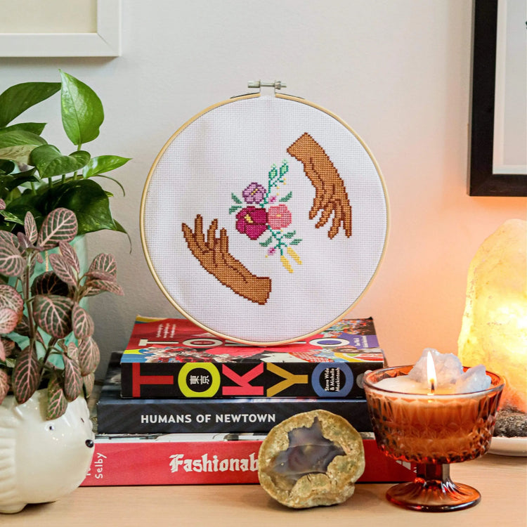 Craft Club Co HANDS HOLD Cross Stitch Kit. The design is shown on a stack of books, with a candle and pot plants around it. The design shows two hands, made from medium brown and dark brown thread, framing a bouquet of flowers. The flowers are light pink, deep pink and purple and have leaves and buds around them.