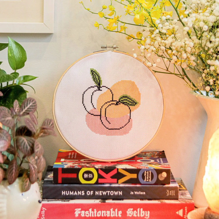 Craft Club Co PEACHY KEEN Cross Stitch Kit. The design shows a line drawing of two peaches. There are organic shapes behind it in orange and pink. There are also organic blotches of olive green behind the leaves on the line art.