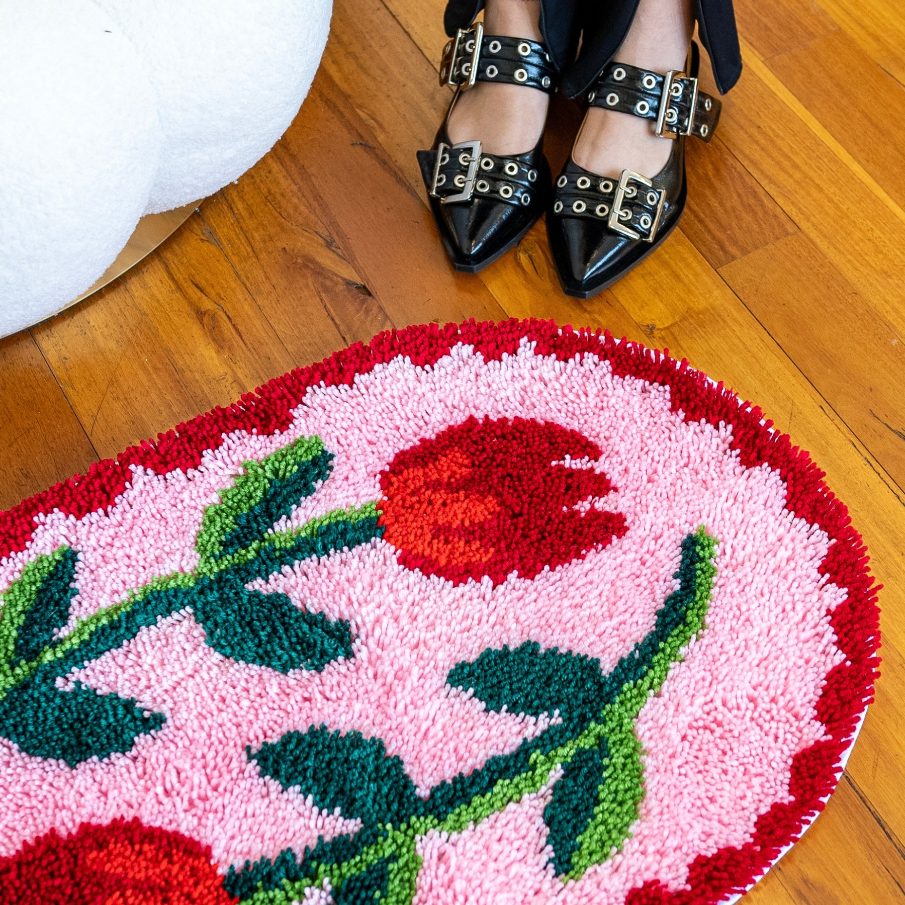 A large latch hooked rug sits on the floor next to a coffee table and chair. The rug has two tulips on it in creamy and brown colours. A persons feet in buckled shoes stands next to the rug.
