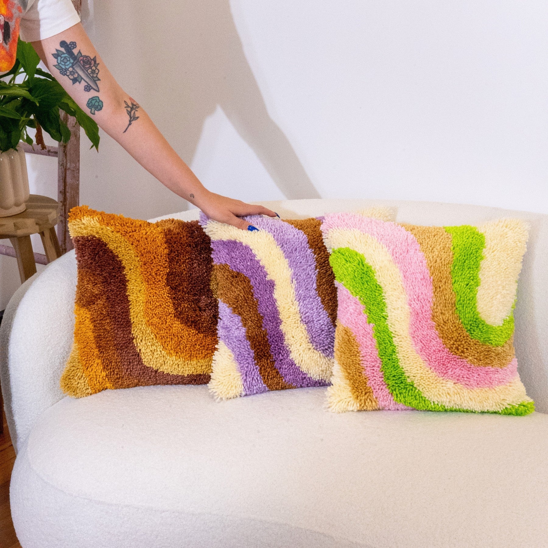 Three latch-hooked cushions sit on a white couch, a hand caresses one of the cushions. The cushion on the left is brown, the one in the middle is purple and the one on the right is green & pink