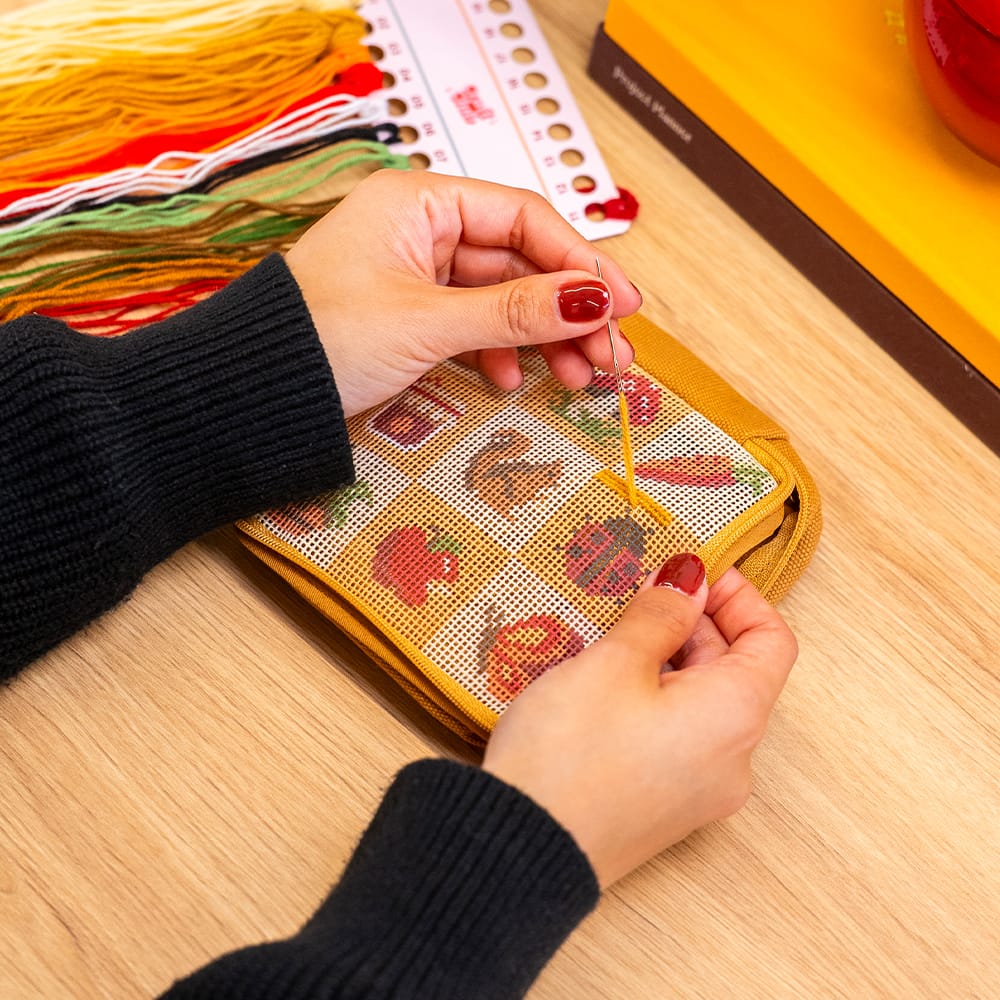Hands stitching the FALL HARVEST CHARMS pouch canvas at a desk beside a thread rack