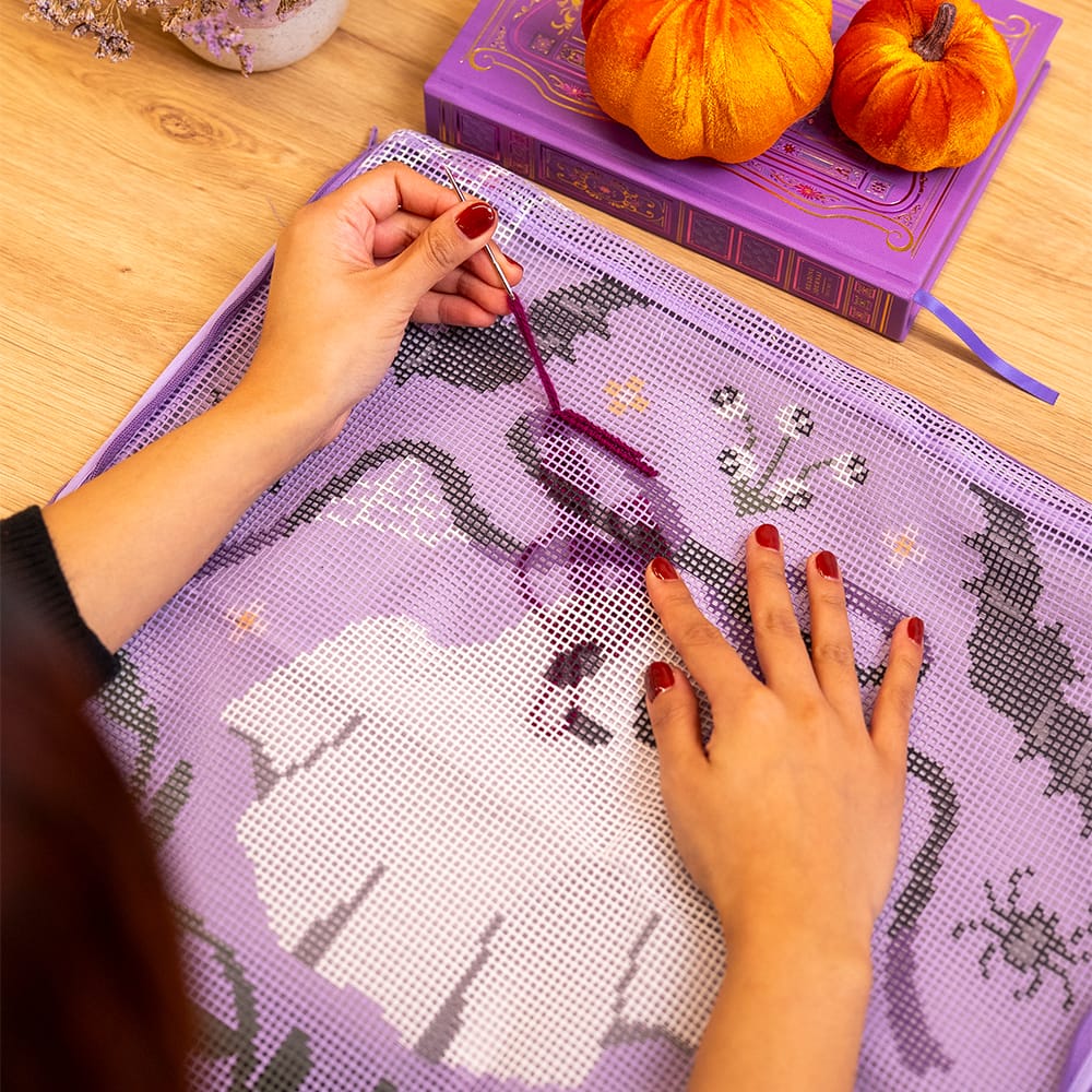 Hands stitching the BOO-TIFUL ghost canvas at a table beside a purple book and velvet pumpkins