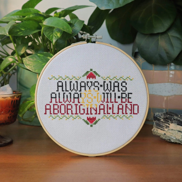 Craft Club Co ALWAYS WAS, ALWAYS WILL BE Cross Stitch Kit. The design shows the text "always was, always will be, aboriginal land" in black, yellow and red yarn. The text has a floral border with wattle and waratah details.