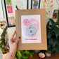 Craft Club Co BEE & BLOSSOM Cross Stitch Kit. A hand holds the kit in its packaging. The kit is packaged in an A4 craft envelope with a blush pink label. The label is a modern style with organic shapes.