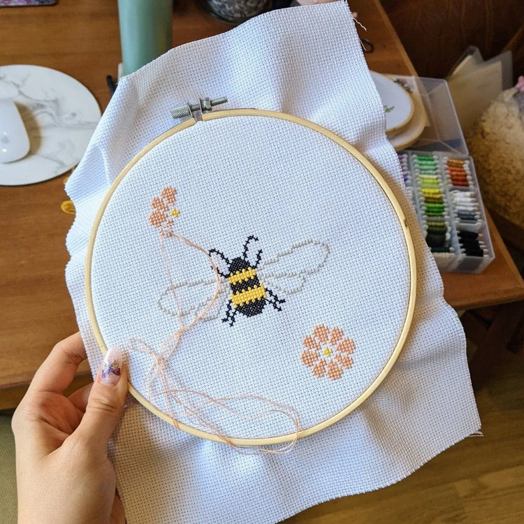 Craft Club Co BEE & BLOSSOM Cross Stitch Kit. The design is mid-creation, with the bee having been completed and two large flowers in the process of being stitched. Light pink thread hangs across the design.