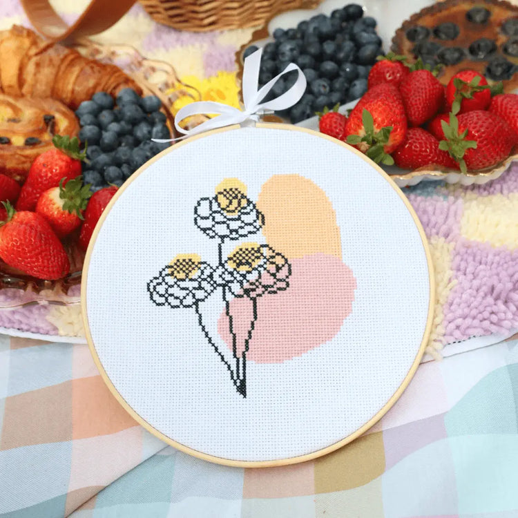 Craft Club Co BLOOM Cross Stitch Kit. The design is three line art flowers, with organic pink and orange shapes behind it. There is also yellow splotches on the flowers. The design sits on a picnic blanket with pastries, blueberries and strawberries in the background.
