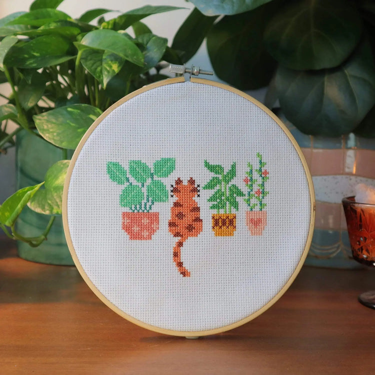 Craft Club Co CAT AMONGST THE POT PLANTS Cross Stitch Kit. The design shows an orange and brown cat sitting amongst three pot plants. One has large leaves with a pink pot, another has tall thin leaves with a brown 70s pot and the third small with pink flowers and a pink pot with a heart.