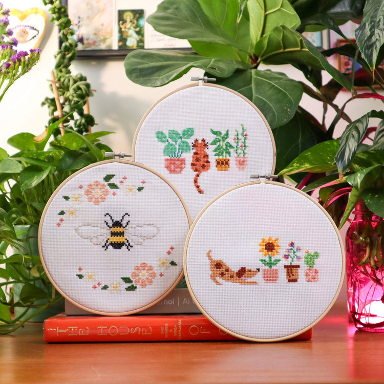 Craft Club Co PUP & THE PLANTS Cross Stitch Kit, Craft Club Co BEE & BLOSSOM Cross Stitch Kit, Craft Club Co CAT AMONGST THE POT PLANTS Cross Stitch Kit. The three designs are shown together in the 'Floral Friends Bundle'. The pup and cat designs match in style and the florals across all pieces work together to create a collection.