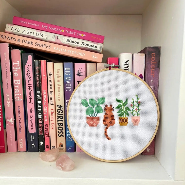 Craft Club Co CAT AMONGST THE POT PLANTS Cross Stitch Kit. The design sits on a bookshelf with all pink books displayed. It has two rose quartz crystals next to it.