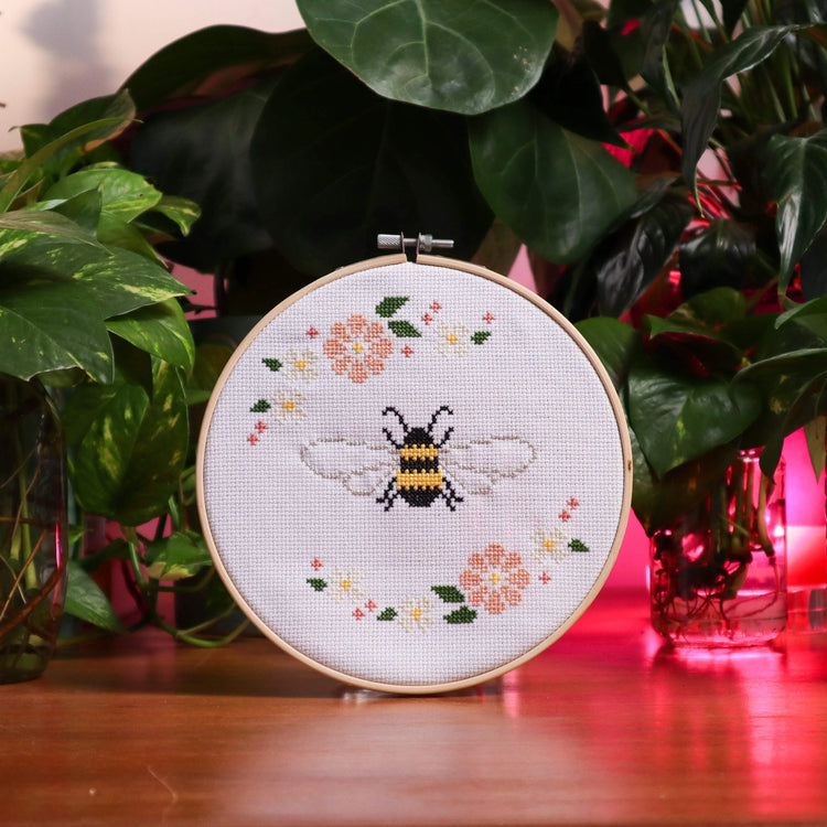 Craft Club Co FLORAL FRIENDS Cross Stitch Kit Bundle. Showing the Bee & Blossom design, it is surrounded by plants and has pink backlighting creating a funky and cool mood.