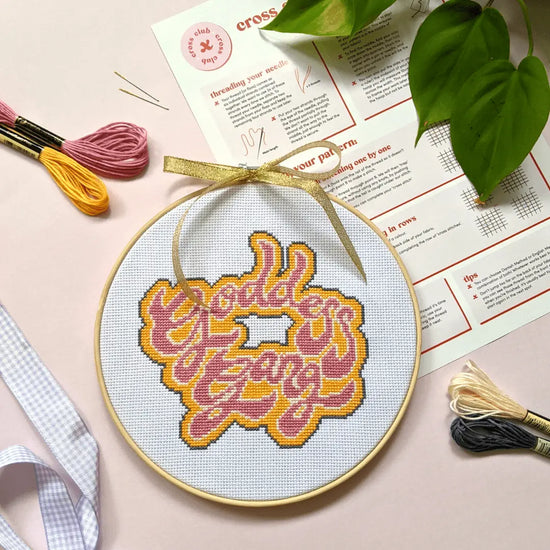   Craft Club Co GODDESS GANG Cross Stitch Kit. The design shows the text "goddess gang" in a large, funky, 70s font. The writing is pink with a cream boarder and the background is orange with a grey boarder. The design sits on a pink background with its instructions and threads surrounding it.