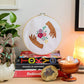 Craft Club Co HANDS HOLD Cross Stitch Kit. The design is shown on a stack of books, with a candle and pot plants around it. The design shows two hands, made from medium brown and dark brown thread, framing a bouquet of flowers. The flowers are light pink, deep pink and purple and have leaves and buds around them.