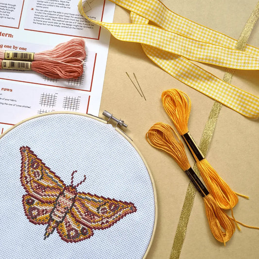 Craft Club Co MOTH Cross Stitch Kit. The design shows a moth with intricate patterns on its wings. The moth is made from warm tones, including maroon, terracotta, deep yellows and pink. The design sits on a yellow background with its thread and ribbons surrounding it.
