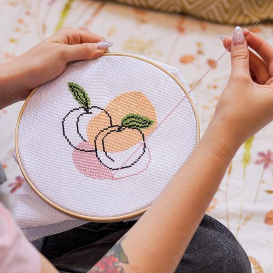 Craft Club Co PEACHY KEEN Cross Stitch Kit. The design is shown mid-creation, with someone stitching the light pink shape behind the piece. They are holding the needle high and a pink thread is visible coming from the design.