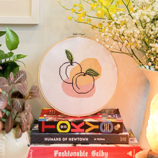 Craft Club Co PEACHY KEEN Cross Stitch Kit. The design shows a line drawing of two peaches. There are organic shapes behind it in orange and pink. There are also organic blotches of olive green behind the leaves on the line art.