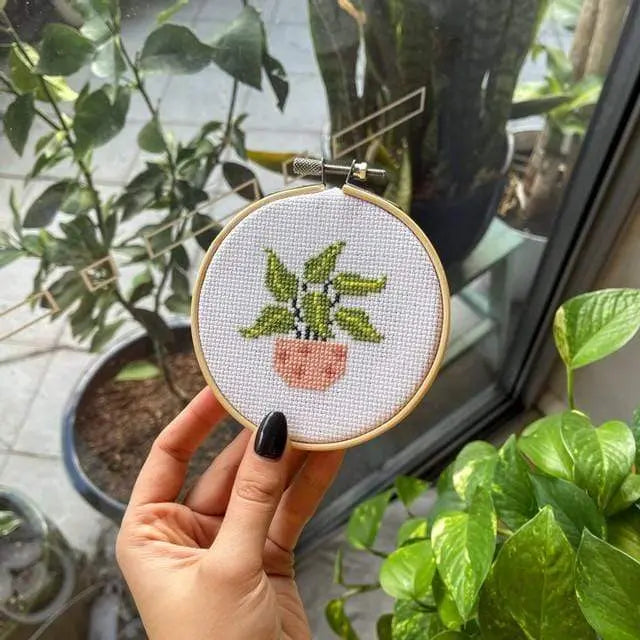 Craft Club Co POT PLANT MINIS Cross Stitch Kit. A hand with black nails holds one of the pot plant designs. There are pot plants in the background.