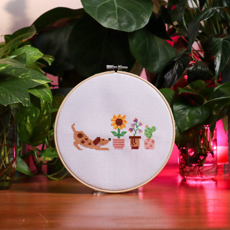Craft Club Co PUP & THE PLANTS Cross Stitch Kit. The design shows a light brown puppy with a wagging tail, stretching amongst three pot plants. One plant is a sunflower in a pink striped pot, the next has purple bell flowers in a brown pot and the last is a cactus in a pink spotted pot.
