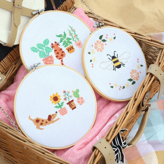 Craft Club Co PUP & THE PLANTS Cross Stitch Kit, Craft Club Co BEE & BLOSSOM Cross Stitch Kit, Craft Club Co CAT AMONGST THE POT PLANTS Cross Stitch Kit. The bundle including the Cat, Pup and Bee designs sits in an open picnic basket on the beach.