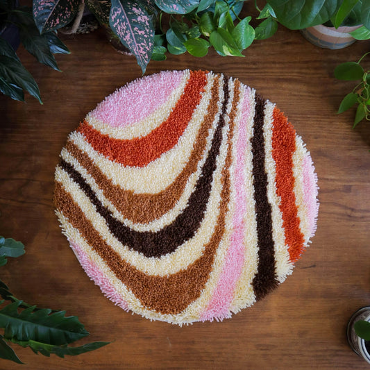 Craft Club Co CARAMEL SWIRL Rug Making Kit. The rug is a round shape, with a 70s style pattern of swirling lines. The background is cream and the lines are pink, orange, light and dark brown.