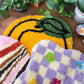 Craft Club Co CARAMEL SWIRL Rug Making Kit, Craft Club Co PEACHY Rug Making Kit, Craft Club Co CHECK ME OUT Rug Making Kit. All three rugs are overlapping on a table surrounded with pot plants and a candle.