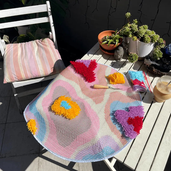 Craft Club Co PSYCHEDELIA Rug Making Kit. The rug is shown only partially done, with small sections of it complete with yarn. A latch-hook and extra yarn sits on the table next to it, along with pot plants and an iced coffee.