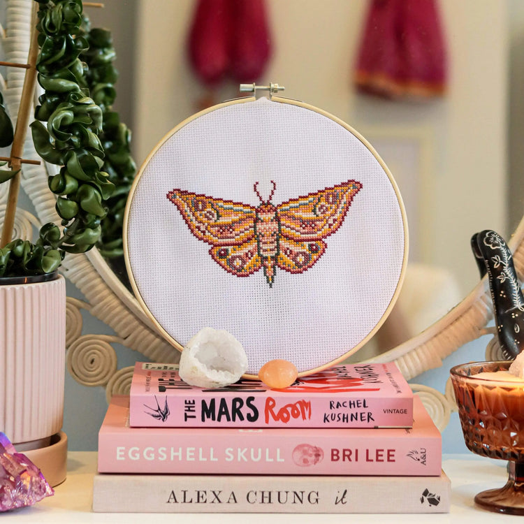 Craft Club Co MOTH Cross Stitch Kit. The design is shown on a stack of books, with a geode and peach selenite crystal next to it.